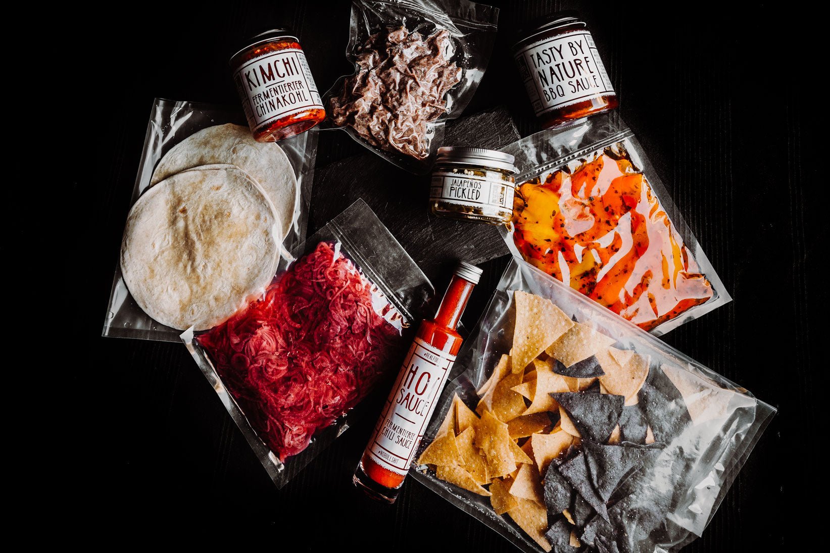 Die Signature Zutaten Wrapstars, wie Tortilla Chips, BBQ Sauce, Pickled Veggies verpackt und auf schwarzem Tisch platziert.