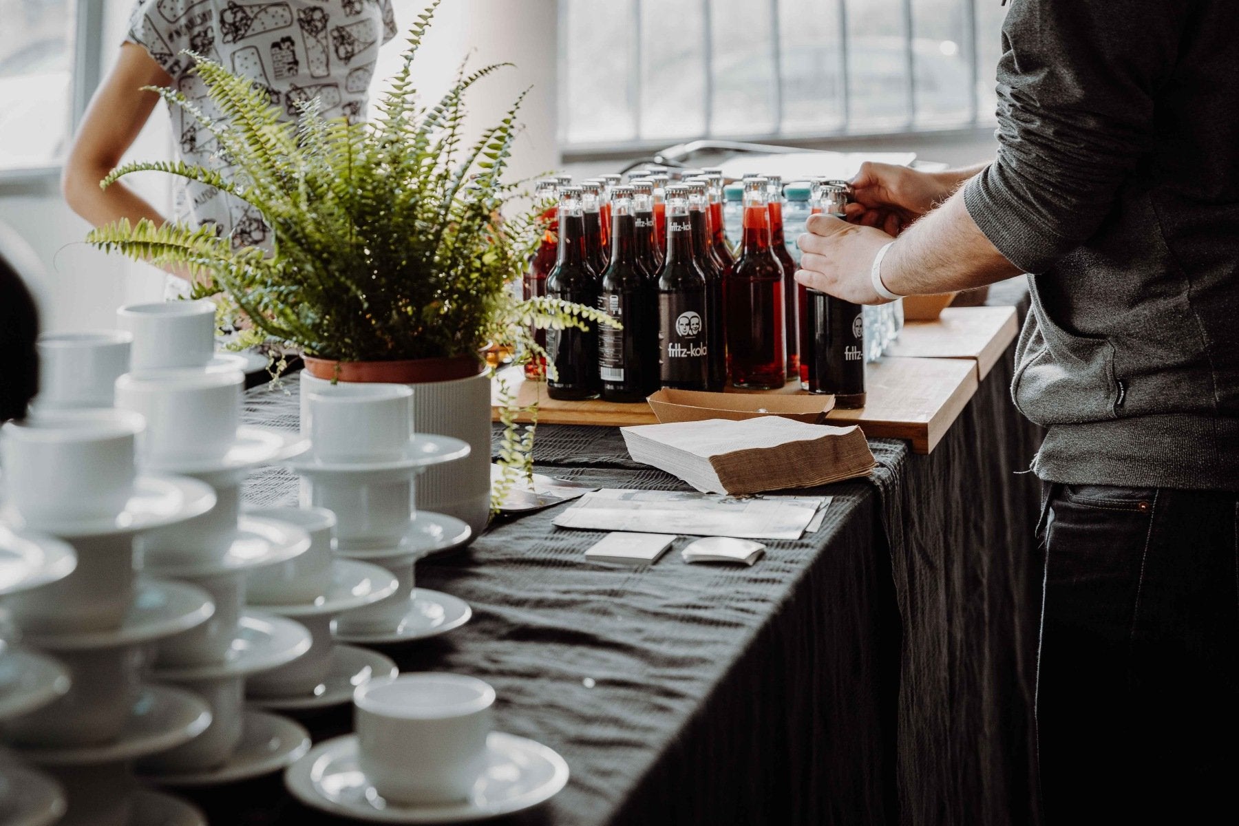 Ein schön dekoriertes Catering auf einem schwarzen Tischtuch mit verschiedenen Limos in Flaschen und gestapelten Kaffeetassen daneben. Eine Person öffnet eine Flasche.